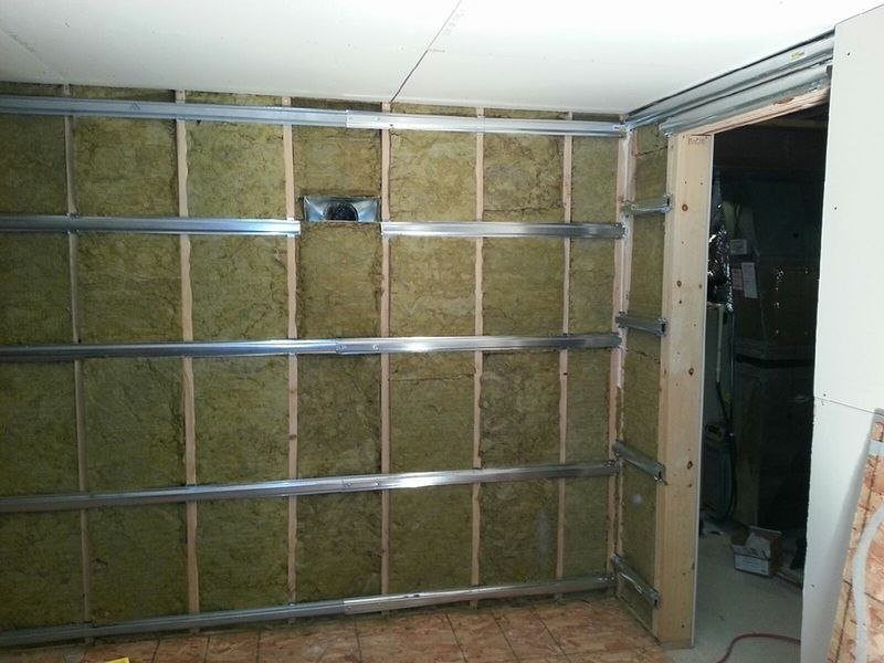 Steel stud framing packed with mineral wool, resilient channel running horizontal across the studs. The channel is the whole trick — it decouples the drywall from the structure so vibration has no rigid path through.